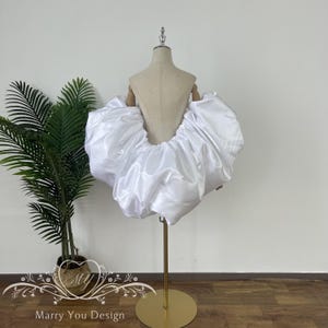 以下が含まれることがあります： 白いサテンのスカートで、ボリューム感のあるギャザーデザイン。マネキンにスカートを展示し、その形と質感を表現しています。この衣服は、おそらく特別な機会のためのファッションコレクションの一部です。