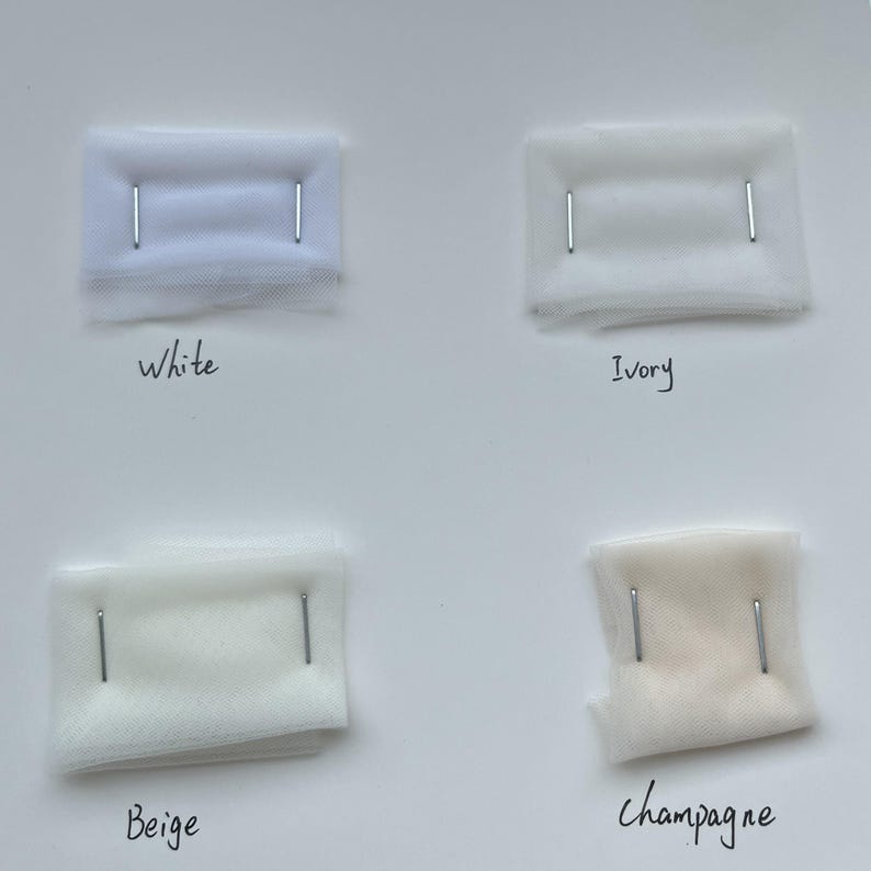 Puede incluir: Cuatro muestras de tela en blanco, marfil, beige y champ&aacute;n, cada una asegurada con dos alfileres plateados. Etiquetas manuscritas identifican cada color. Las muestras est&aacute;n dispuestas sobre una superficie blanca, mostrando los diferentes tonos.