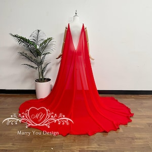 May include: A vibrant red cape with a long train, draped over a mannequin. The cape features a deep V-neck design and is made of a flowing, lightweight fabric. The image includes the text "Marry You Design" within a heart-shaped logo.