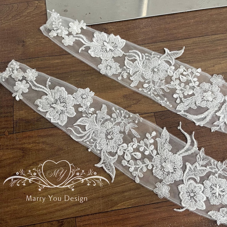 Puede incluir: Dos tiras de encaje floral blanco con intrincados dise&ntilde;os bordados de flores y hojas. El encaje est&aacute; sobre una superficie de madera. El texto "Marry You Design" es visible.