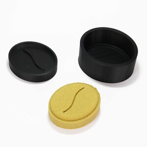 May include: A coffee bean-shaped mold with a matching lid and a separate black container. The mold is tan with a curved groove, and the lid and container are black. The items are arranged on a white surface.