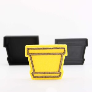 May include: A yellow flower pot with a brown outline. The pot is sitting on a white surface.