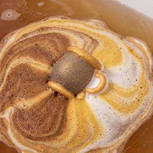 Puede incluir: Una bomba de baño con forma de taza flota en el agua, creando patrones de espuma en espiral de color marrón, dorado y blanco. La bomba de baño es de color marrón oscuro con un borde y asa dorados metálicos, disolviéndose en el líquido.