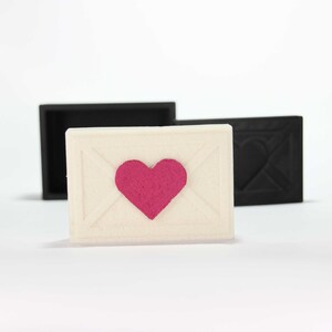 May include: A white envelope with a pink heart in the center. The envelope is sitting on a white surface.