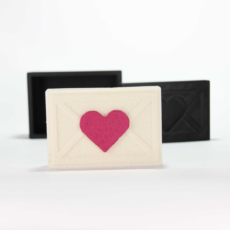 May include: A white envelope with a pink heart in the center. The envelope is sitting on a white surface.