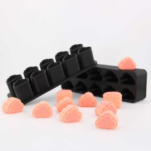 May include: Two black silicone molds for making candy or soap. One mold is shaped like a cloud and has 12 cavities. The other mold is shaped like a heart and has 8 cavities. There are several pink cloud-shaped candies on a white surface.