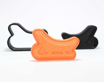 Könnte beinhalten: Orangener Plastik-Ausstecher in Form eines Schlittens. Der Ausstecher liegt auf einer weißen Oberfläche neben einem schwarzen Plastik-Ausstecher in der gleichen Form.