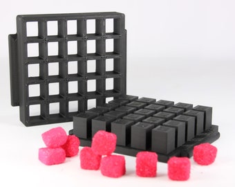 Op de afbeelding: Een zwarte plastic mal met een rasterpatroon, gevuld met kleine zwarte blokjes. Roze suikerklontjes zijn verspreid rond de mal.