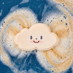 May include: A pale cream-colored cloud-shaped bath bomb with a simple printed face featuring two dark oval eyes, a small dot nose, and a curved mouth, floating in deep blue water surrounded by swirling white and tan foam bubbles.