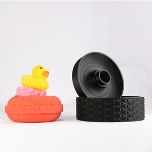 May include: An orange bath bomb with a yellow rubber duck and pink foam, next to a black bath bomb mold. The mold consists of two parts, a cylinder and a patterned base. The items are on a white background.