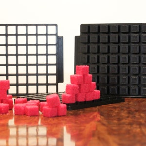 May include: Two black plastic grids with a stack of pink sugar cubes on one of the grids. The sugar cubes are arranged in a pyramid shape.