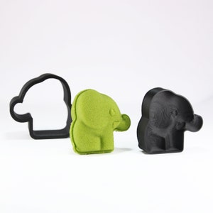 Puede incluir: Tres elementos con forma de elefante. Un cortador de galletas negro, un elefante verde difuso y un elefante negro impreso en 3D. El elefante verde tiene ojos y orejas visibles. El cortador de galletas es un simple contorno de la forma del elefante.