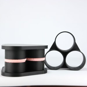 May include: A black plastic 3D printed object with three circular openings. The object is designed to hold three cylindrical items. The object is sitting on a white surface.