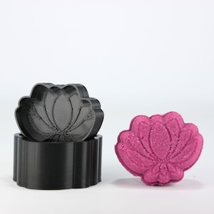 Puede incluir: Un molde negro con forma de flor de loto y una bomba de baño rosa terminada. El molde está hecho de plástico y tiene un diseño detallado de flor de loto. La bomba de baño es de color rosa vibrante y tiene una forma similar a la flor de loto.