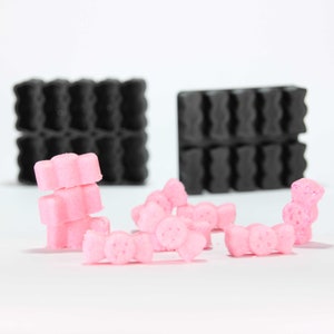 May include: Pink sugar candies shaped like bows and flowers, stacked and scattered on a white surface. Two black silicone molds are in the background.