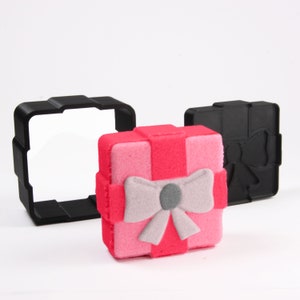 Puede incluir: Una caja de regalo rosa con un lazo gris y un molde de plástico negro de una caja de regalo.