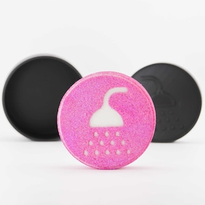 Könnte beinhalten: Eine pinkfarbene, kreisförmige Badebombe mit einem weißen Duschkopf-Design, flankiert von zwei schwarzen, kreisförmigen Objekten. Die Badebombe hat eine glitzernde Textur.
