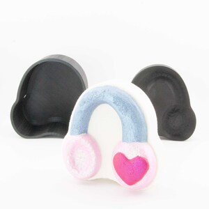 Pode incluir: Uma bomba de banho branca em forma de coração em forma de fones de ouvido, com fones de ouvido rosa e uma faixa de cabeça azul. Um coração rosa está em um fone de ouvido. Dois moldes pretos estão no fundo.