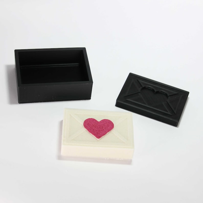 May include: A white box shaped like an envelope with a pink heart in the center. The box is open and the lid is black with a heart-shaped cutout.