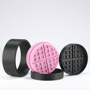 May include: Three 3D printed waffle molds, one pink and two black. The molds are circular and have a waffle pattern.