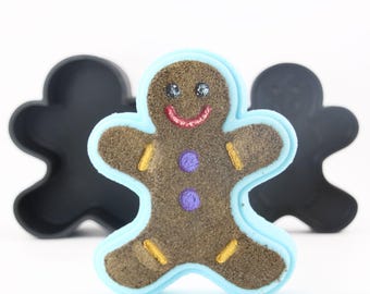 Puede incluir: Un molde para galletas de hombre de jengibre con un contorno azul. El molde está hecho de un material marrón similar a la arena y tiene una cara sonriente con ojos negros, una boca roja y dos botones morados. El molde está rodeado por dos moldes para galletas de hombre de jengibre negros.