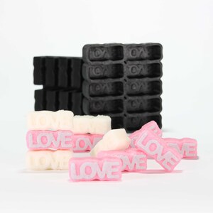 May include: A close-up of pink and white sugar cubes with the word "LOVE" embossed on them. The sugar cubes are arranged in a pile on a white surface. There is a black plastic mold in the background with the word "LOVE" embossed on each of the squares.