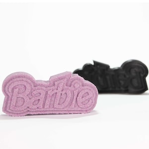 Pode incluir: Dois logotipos "Barbie" impressos em 3D, um rosa e outro preto. O logotipo rosa está em foco e o logotipo preto está desfocado no fundo.