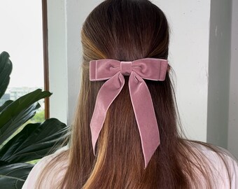 May include: A dusty rose velvet hair bow with a large, tied bow. The bow is worn in the hair of a person with long, brown hair.