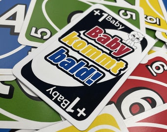 May include: A black playing card featuring the text Baby kommt bald! in red, yellow, and blue, with a small baby icon. The card is surrounded by a scattered pile of colorful numbered playing cards in red, blue, green, and yellow hues.