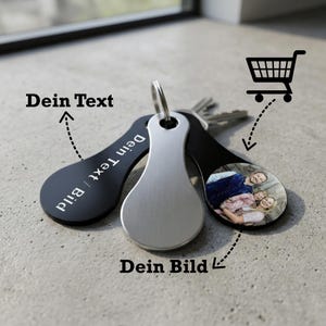 Könnte beinhalten: Drei tropfenförmige Schlüsselanhänger an einem Metallring. Einer ist aus gebürstetem Silber, einer ist schwarz mit dem Text "Dein Text / Bild" und einer ist schwarz mit einem kreisförmigen Farbfoto von zwei Personen. Inklusive Einkaufswagen-Symbol.