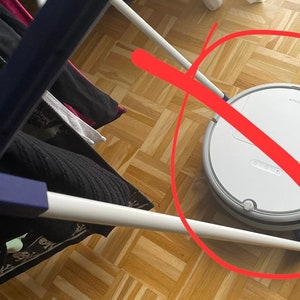 以下が含まれることがあります： 丸い形をした白いロボット掃除機で、グレーの縁取りがあります。掃除機は木製の床にあり、背景には青と白の洋服ラックがあります。