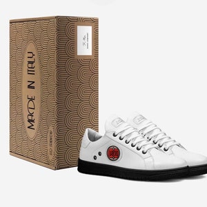 May include: White leather sneakers with black soles and laces, next to a cardboard box. The box has a repeating arch design and the words "MADE IN ITALY". The sneakers have a red and orange circular logo that says "LUVE".