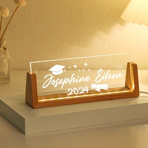 May include: A wooden base with a clear acrylic sign that reads "Josephine Eileen 2024" with a graduation cap and diploma graphic. The sign is illuminated with warm white LED lights.