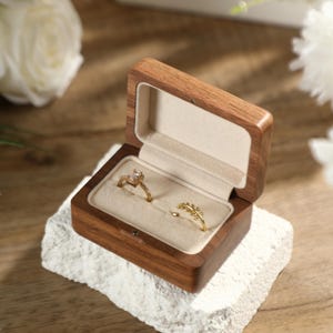 Könnte beinhalten: Geöffnete Holz-Ringbox mit zwei goldenen Ringen. Die Box steht auf einer weißen, strukturierten Oberfläche, mit weißen Blumen im Hintergrund. Ein Ring hat einen großen, klaren Stein, der andere ein zartes Blattdesign. Die Box ist aus dunklem Holz.
