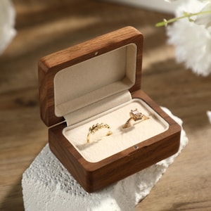 Könnte beinhalten: Geöffnete Holzringbox mit zwei goldenen Ringen. Ein Ring hat einen quadratischen Edelstein, der andere ein zartes Blattdesign. Die Box ist mit cremefarbenem Stoff ausgekleidet und steht auf einer weißen, strukturierten Oberfläche.