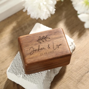 以下が含まれることがあります： 「Jochen & Lya」と日付「20.08.2025」が刻まれた、ヒンジ付きの蓋が付いた木製の箱。箱は白いテクスチャのある表面に置かれ、背景には白い花があります。箱は天然の木目仕上げです。