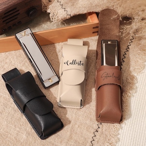 May include: Three harmonica cases with personalized names. The cases are made of brown, black, and beige leather. The harmonicas are silver and have a rectangular shape.