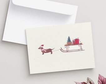 May include: A cream-colored greeting card featuring a watercolor illustration of a brown dachshund wearing a red and white Santa hat and scarf, pulling a wooden sled loaded with a small green Christmas tree and red gift boxes with bows.