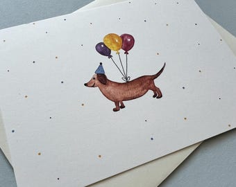 May include: A watercolor birthday card featuring a brown dachshund wearing a party hat and holding three balloons. The balloons are yellow, purple, and maroon. The card is on a white background with small dots and comes with a cream-colored envelope.
