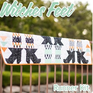May include: A Halloween-themed runner kit with the text "Witches Feet" and "Runner Kit". The runner features a repeating pattern of witch's feet and other Halloween-themed designs in black, orange, and white. The runner is displayed on a brown metal railing.