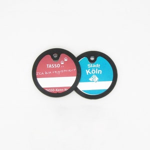Könnte beinhalten: Zwei runde schwarze Hundemarken mit weißem Text. Die Marke links ist rot und sagt "TASSO e.V. Ich bin registriert TASSO-Kenn-Nr." Die Marke rechts ist blau und sagt "Stadt Köln".