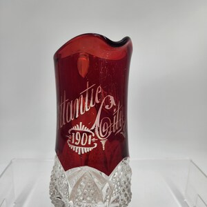 May include: A red glass pitcher with a clear glass base. The pitcher has a wavy rim and is decorated with white lettering that reads "Atlantic City 1901".