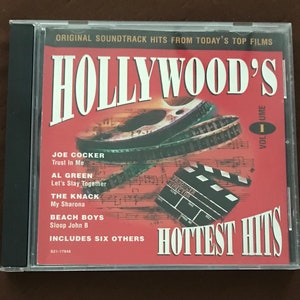 May include: A CD case for "Hollywood's Hottest Hits, Volume 1." The cover is red with the title in large white letters. It features film reels, a clapperboard, and a list of songs by Joe Cocker, Al Green, The Knack, and Beach Boys.