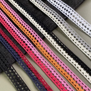 May include: Assortment of seven different lace trims in various colours, including black, navy, red, pink, orange, white and grey. The trims are arranged in a row on a dark blue denim fabric background.