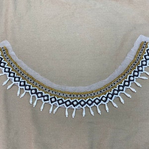 May include: A decorative collar with a curved shape, featuring a detailed beaded design. The collar has a white mesh base and is adorned with black, gold, and white beads, including small pearl-like accents and dangling teardrop beads.