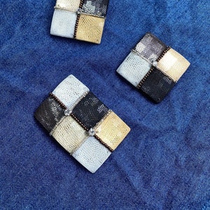 May include: Three decorative square-shaped brooches with a patchwork design. The brooches are made of beige, white, and black leather with a snakeskin pattern. Each brooch has a small clear rhinestone in the center.