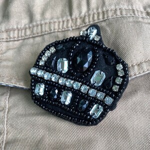 Puede incluir: Una corona de fieltro negro con un borde de cuentas y diamantes de imitación transparentes y negros.