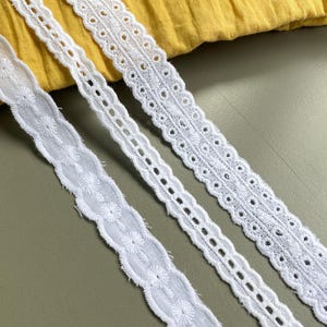 May include: Three white cotton lace trims with different patterns. The lace trims are arranged horizontally on a green surface.