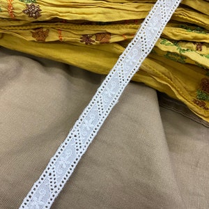 May include: White cotton lace trim with a delicate floral pattern. The lace is shown on top of a yellow fabric with embroidery.