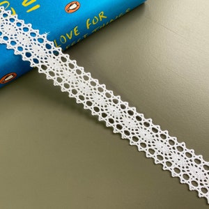 May include: White cotton lace trim with a delicate, intricate pattern. The lace is approximately 1 inch wide and has a scalloped edge.
